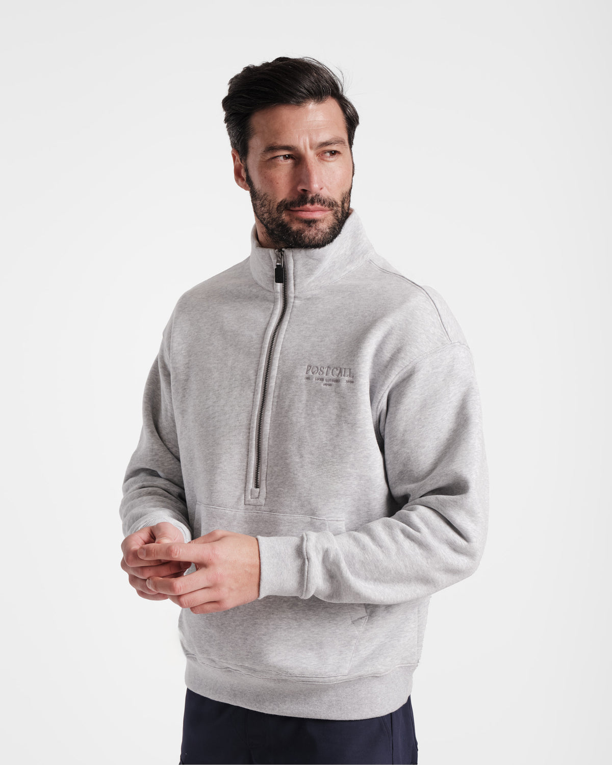 Man wearing heather grey half zip sweatshirt with a kangaroo pocket, tonal Post Call logo embroidered on the left chest, and leather zip pull zipped to the top.