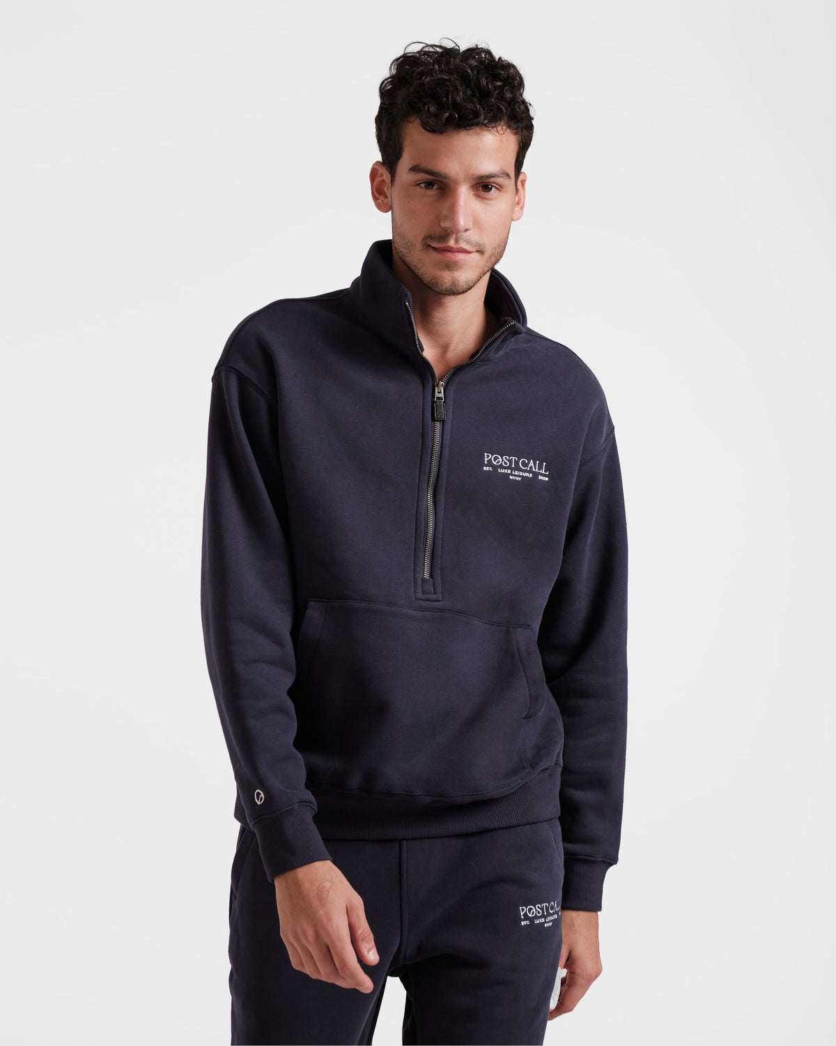 Man wearing navy half zip sweatshirt with kangaroo pocket and white Post Call embroidered logo on the left chest.