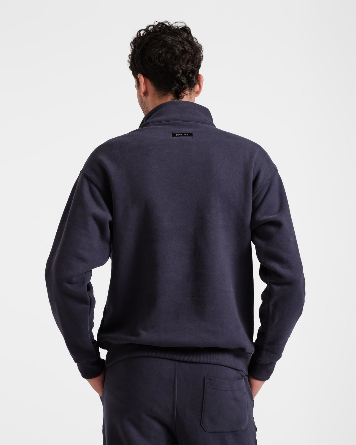 Back side view of man wearing navy half zip sweatshirt with black Post Call fabric label sewn under the neck seam on the back panel.