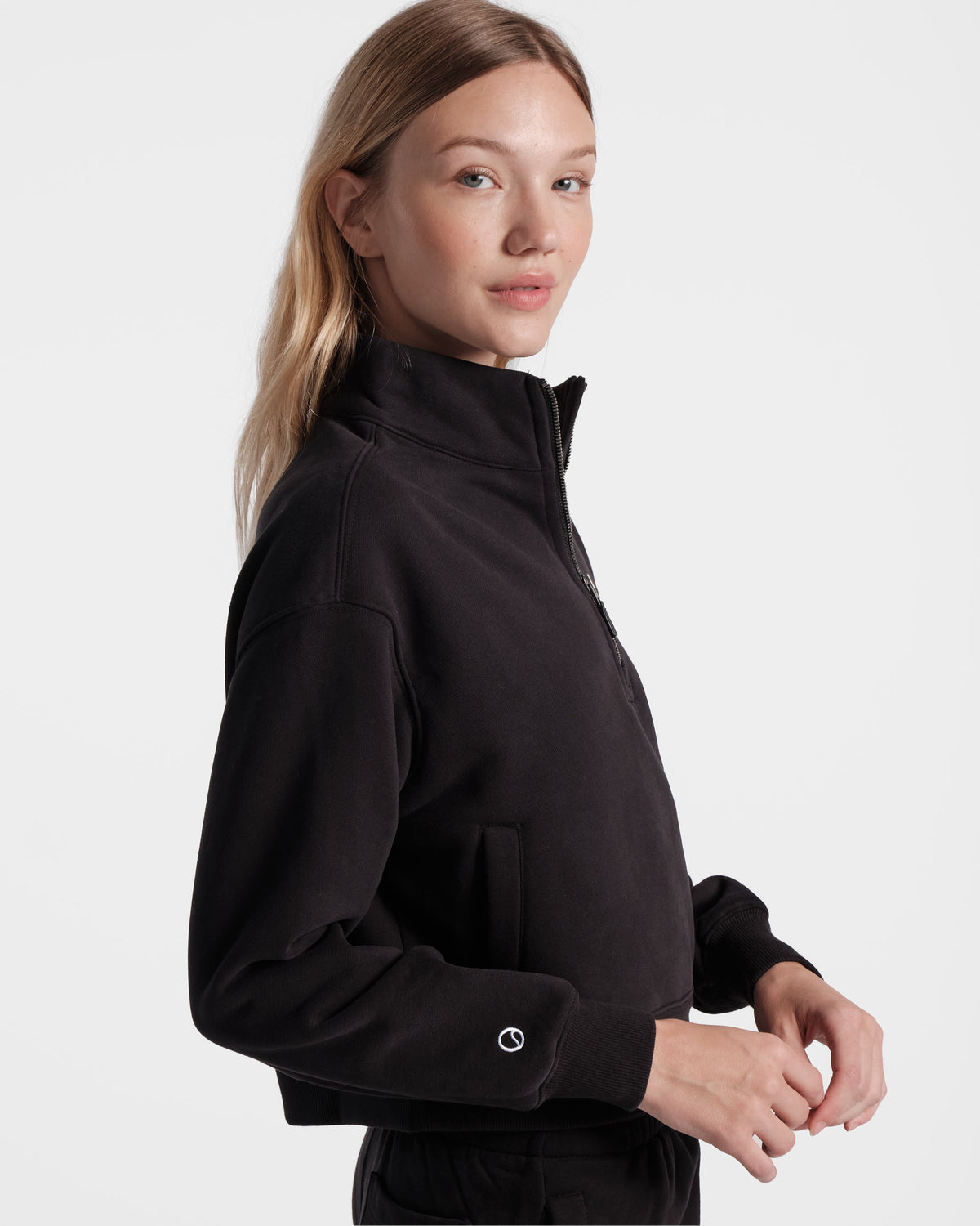 Side view of woman wearing black half zip sweatshirt with the white embroidered small logo emblem above the wrist cuff.