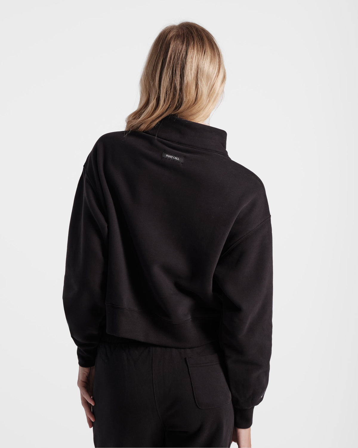 Back side view of woman wearing black half zip sweatshirt with a black Post Call fabric label sewn under the collar on the back panel.
