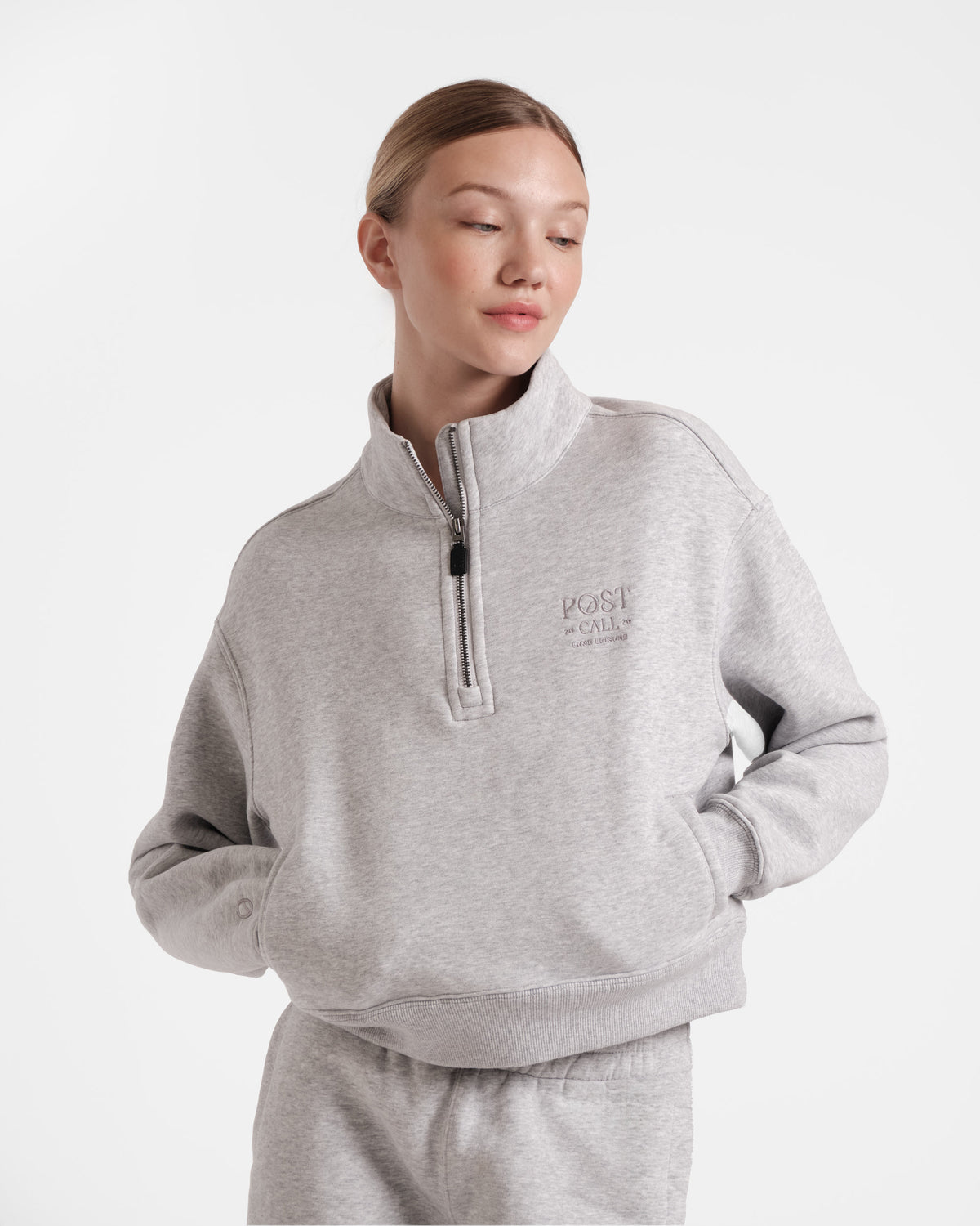Woman wearing heather grey half zip sweatshirt with a leather zip pull and tonal Post Call logo embroidery on the left chest with both hands in her sweatshirt pockets.