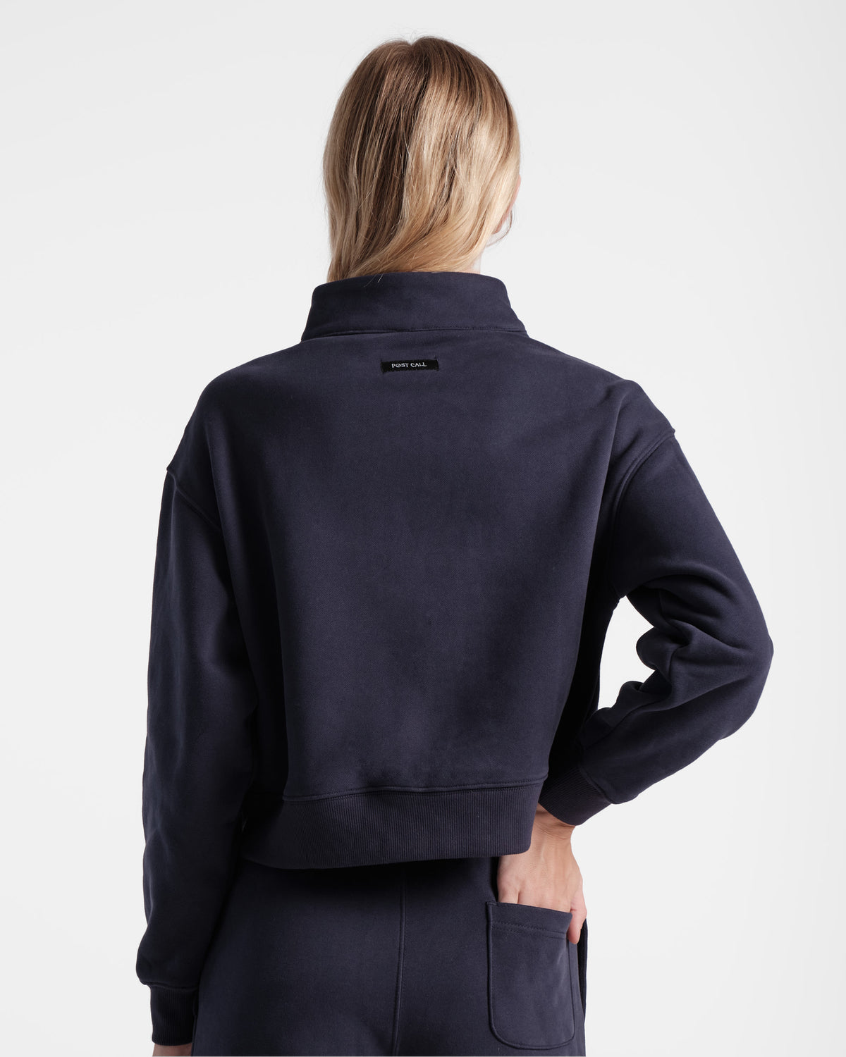Back side view of woman wearing navy half zip sweatshirt with black Post Call fabric label sewn under the neck seam on the back panel. 