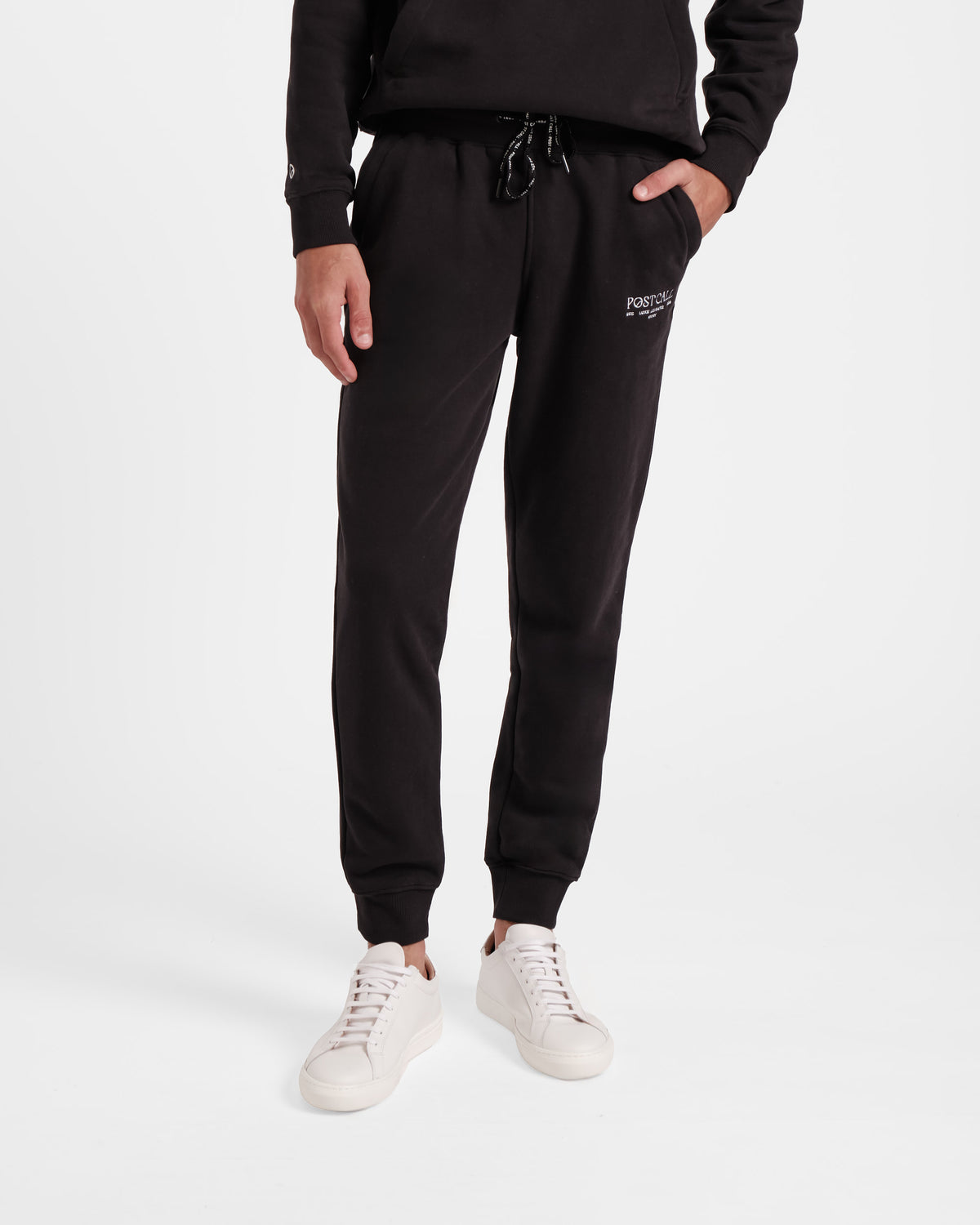 Waist down view of man wearing black jogger sweatpants with stretch ankle cuffs, black contrast branded waist tie, and white embroidered Post Call logo on the thigh.