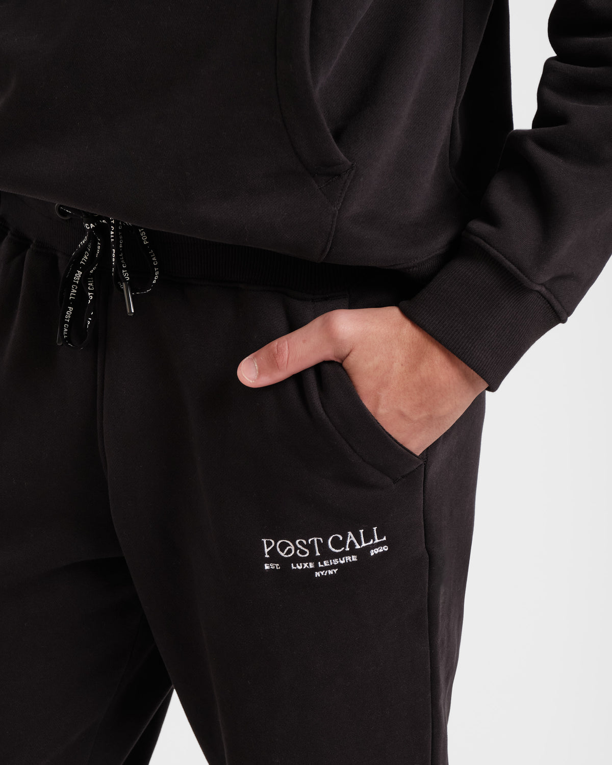 Close up of man's hand in black jogger sweatpant pocket with white embroidered Post Call logo and black contrast branded waist tie.