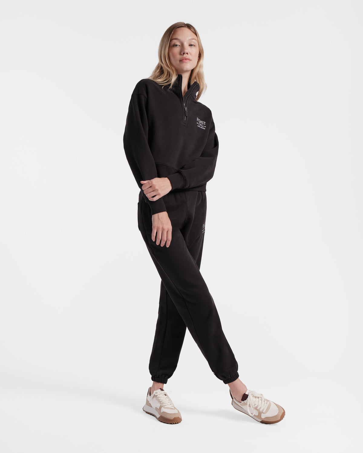 Model wearing women's black jogger sweatpants with a matching half zip sweatshirt standing with her legs crossed.