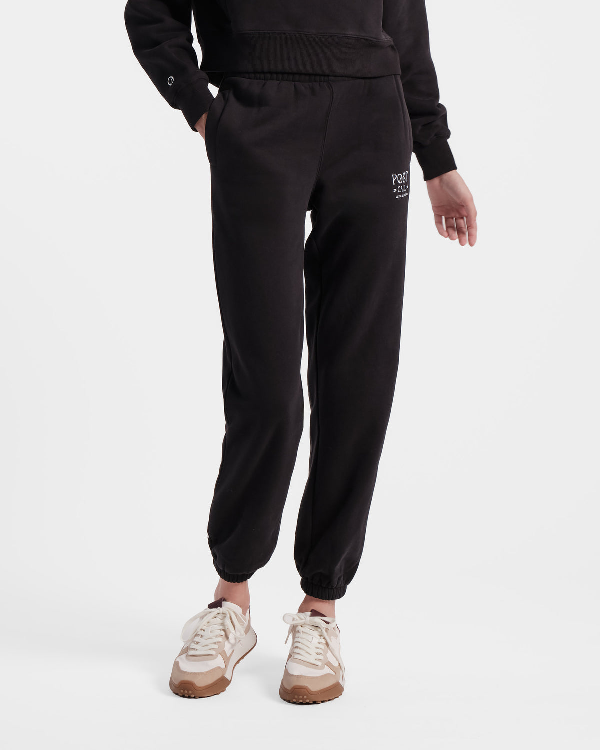 Woman wearing black jogger sweatpants with elasticated ankle cuffs and contrast Post Call logo embroidery in white on the upper thigh.