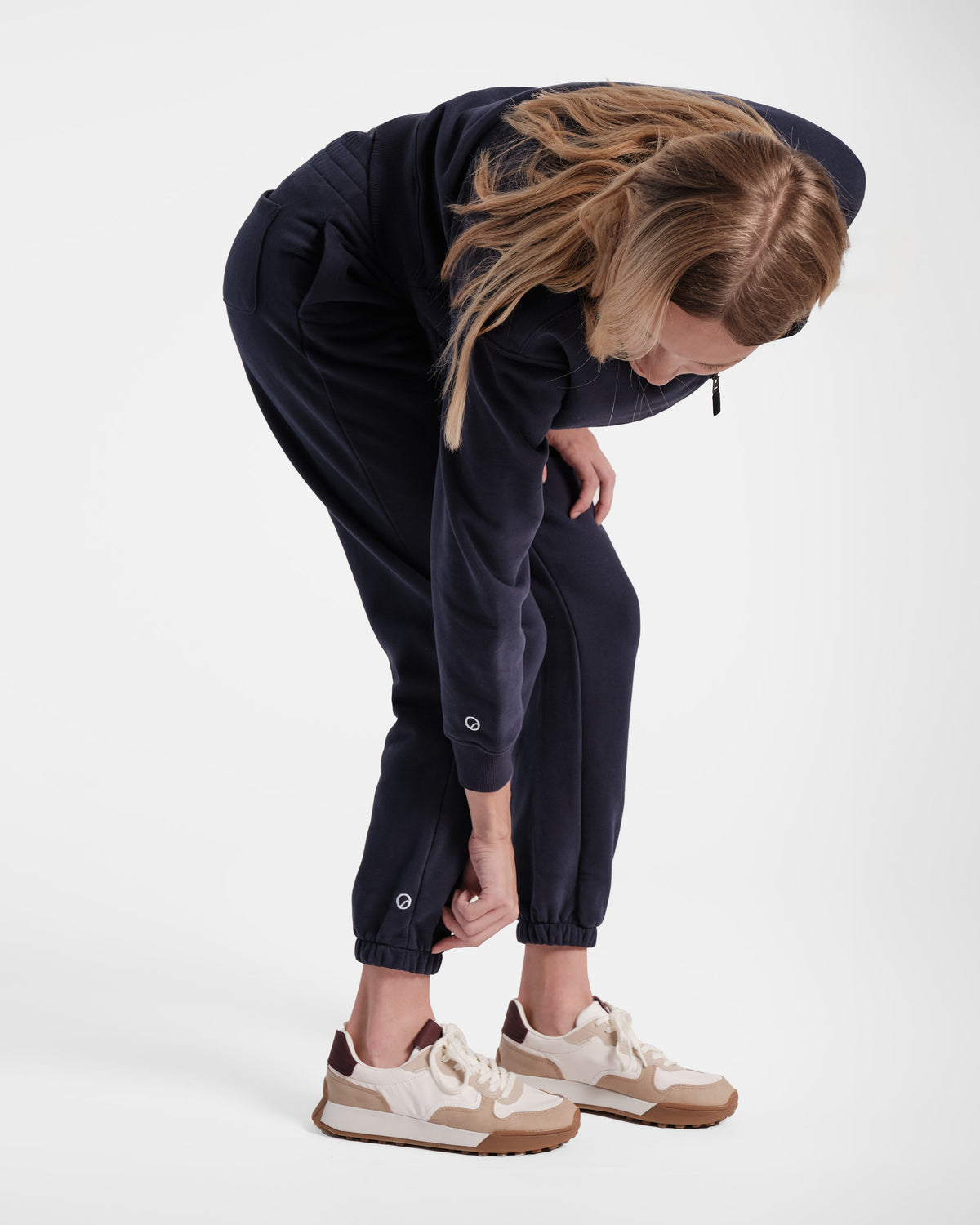 Woman bending over adjusting her ankle cuff on her navy jogger sweatpants with small logo contrast embroidery above the cuff of the pants and matching sweatshirt.