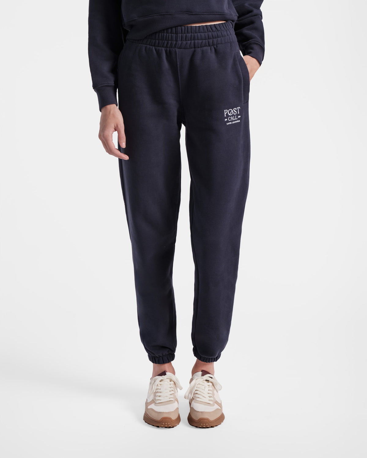 Woman wearing navy jogger sweatpants with elasticated ankle cuffs and contrast Post Call logo embroidery on the upper thigh.