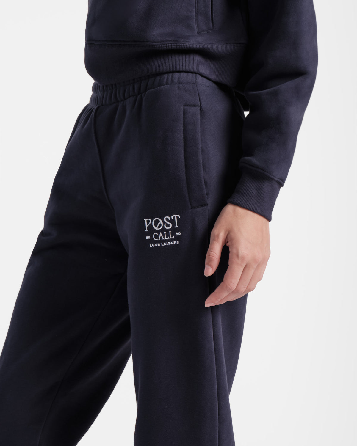 Close up angled view of the side of woman in navy jogger sweatpants with contrast logo embroidery and side pocket .
