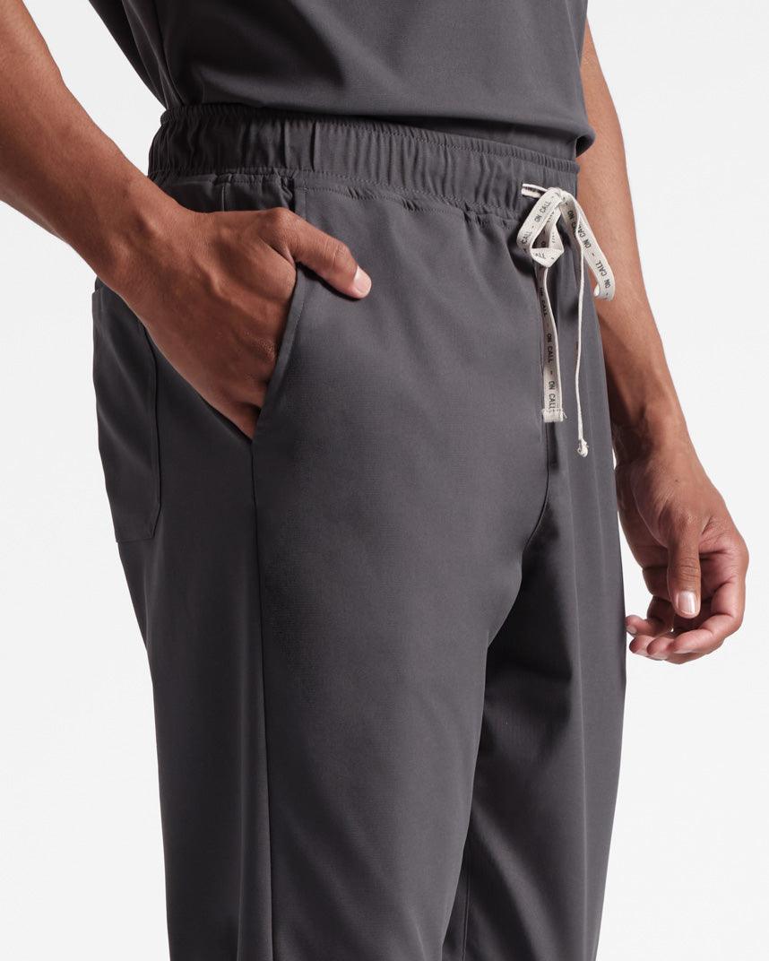 Close up view of man in dark grey scrub pants, focusing on the branded drawstring and waist pocket with his hand inside.