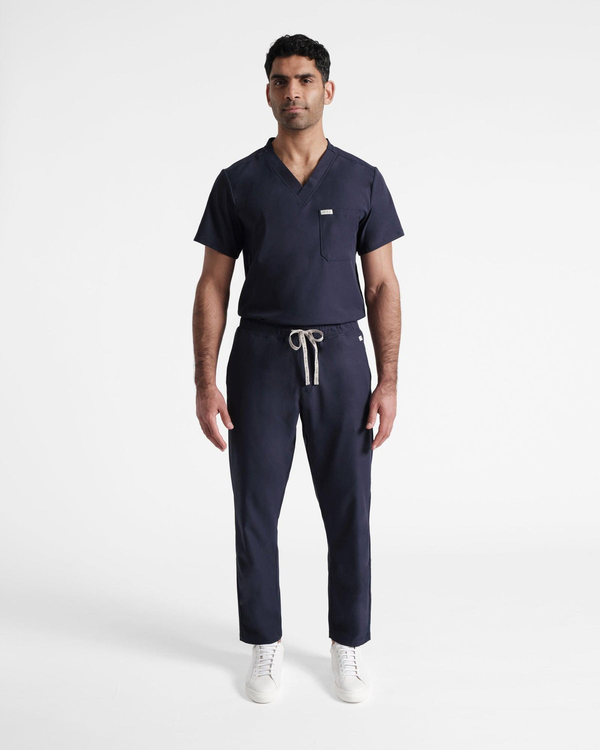 Man in a navy v-neck scrub shirt tucked into matching straight leg flat front scrub pants. 