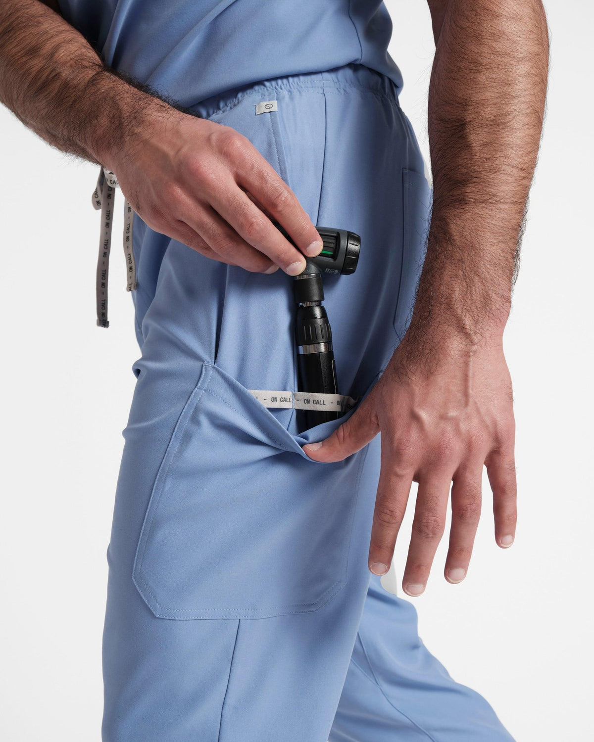 Side view of man pulling down the side leg pocket on his light blue cargo scrub, revealing an Otoscope secured in an On Call branded elastic loop.