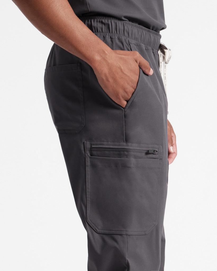 Man in dark grey cargo scrub pants, zoomed in on the waist to the knees with his hand in the waist pocket.