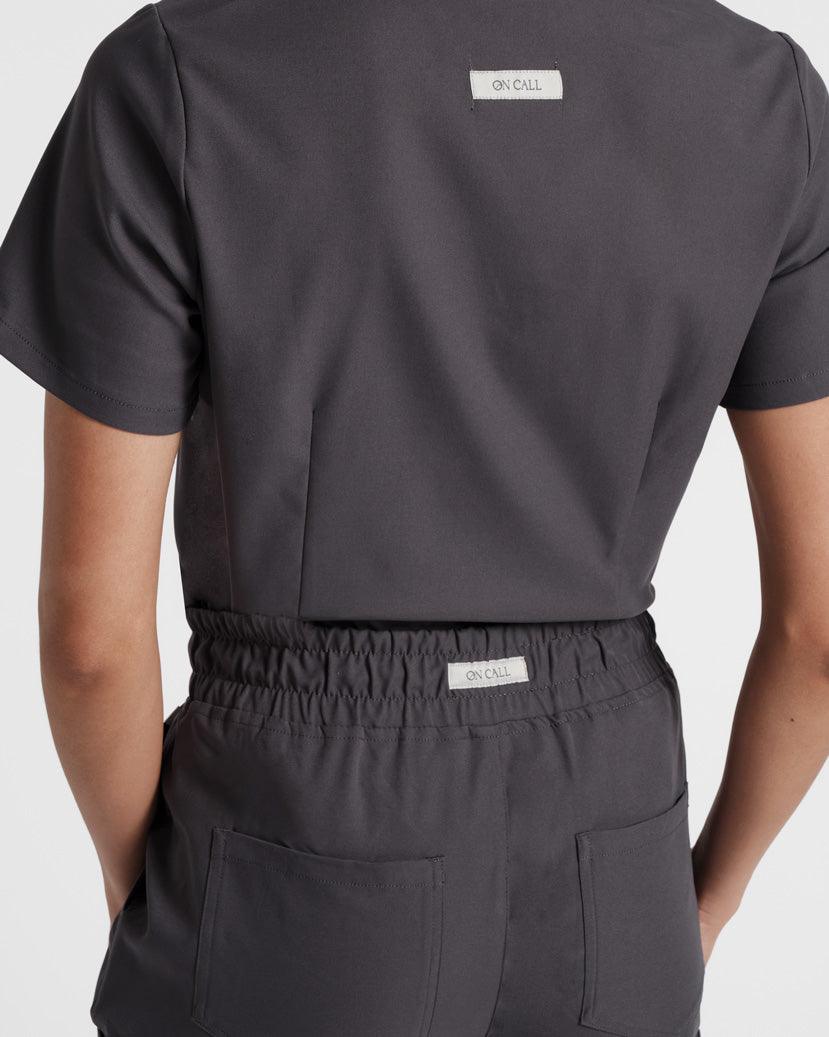 Close up back side view of woman in a matching scrub top and pant set in dark grey with darting on the top.