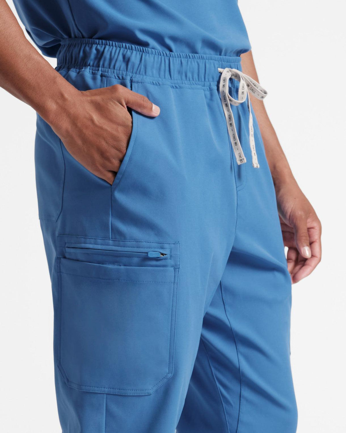Close up angled view of man in bright blue cargo scrub pants, focusing on large side leg pocket and his hand in the waist pocket.