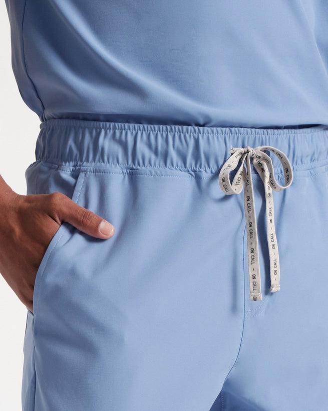 Close up view of man in flat front light blue scrub pants, focused on hand in pocket and branded drawstring at the waist.