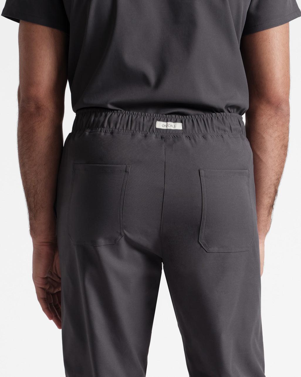 Close up back side view of man wearing dark grey scrub pants with two back pockets and an On Call logo fabric label sewn into the waistband.