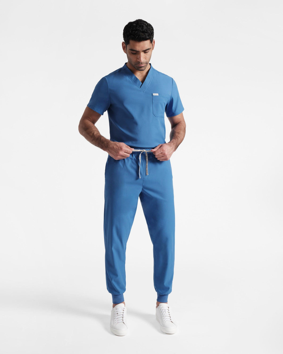 Man wearing bright blue jogger scrub pants with a matching v-neck top tucked in, tying the drawstring at his waist.