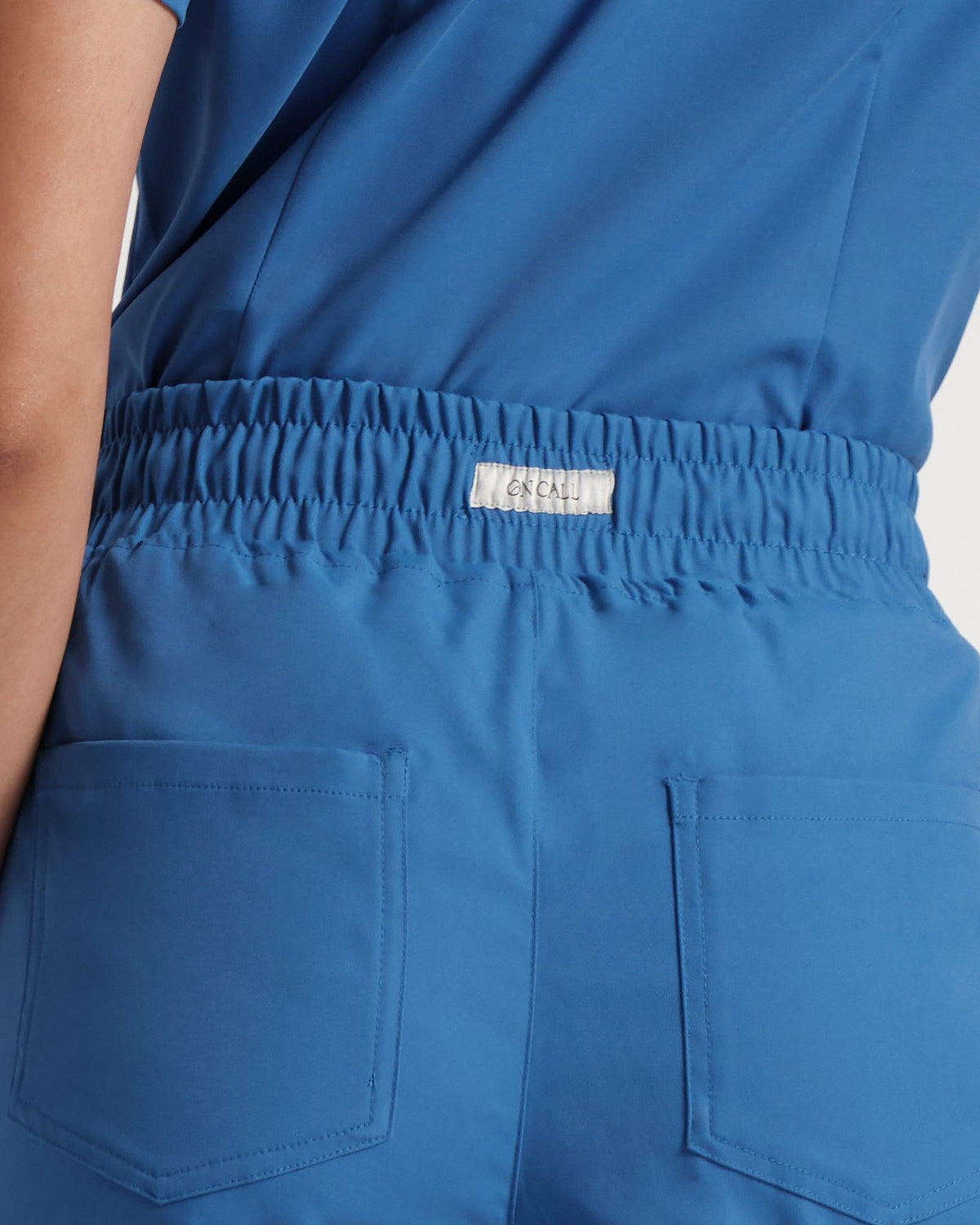 Back side view of woman in bright blue scrub pants and matching top tucked in, zoomed into the waistband with a logo label sewn into the outside.