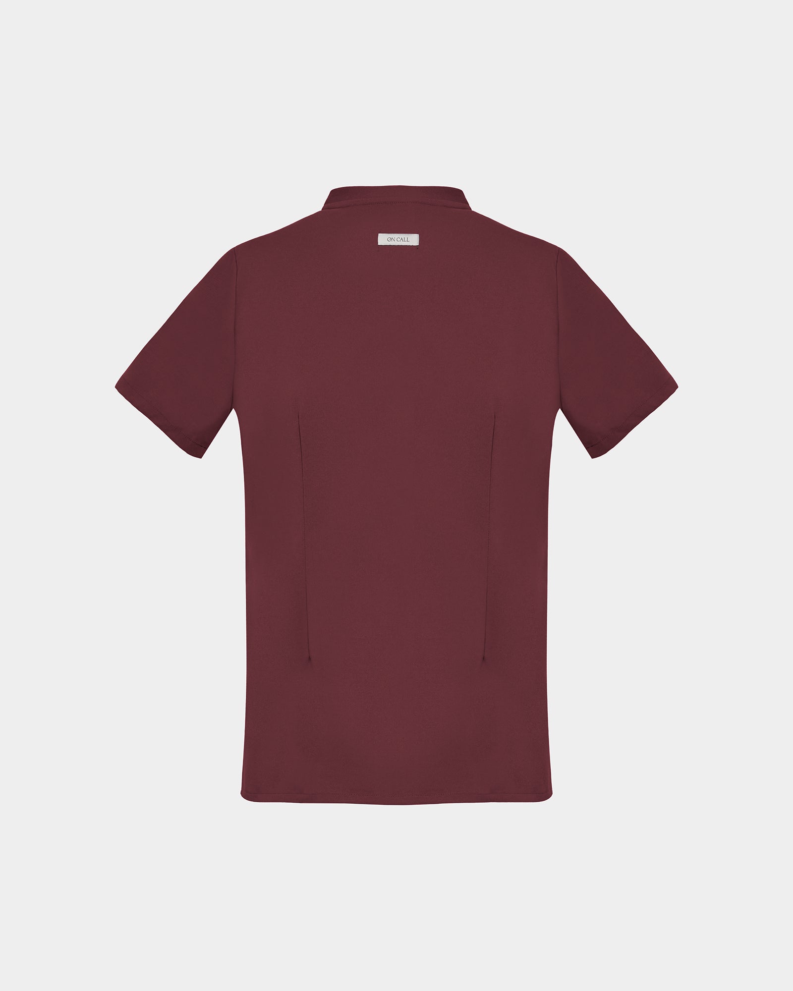 A short-sleeve scrub shirt in a deep heme red color, shown from the back, featuring two subtle darts, against a light gray background.