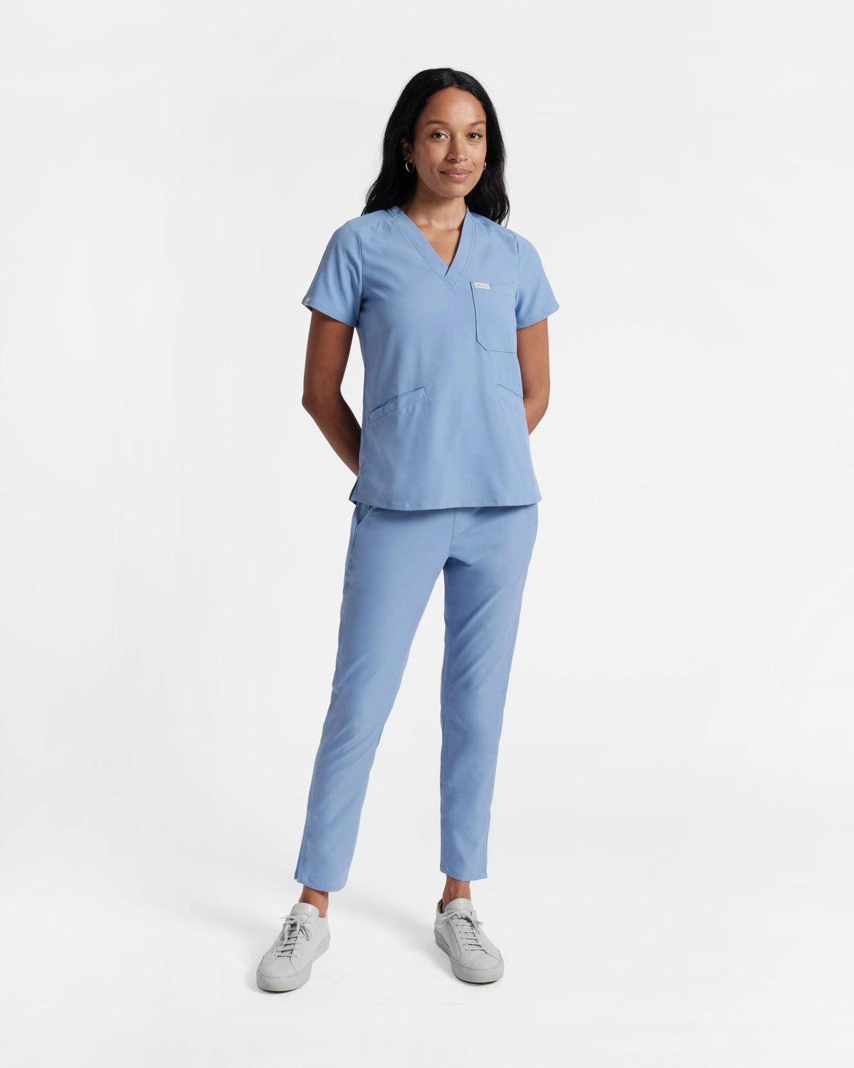 Woman in a v-neck light blue scrub shirt with a chest pocket and two front pockets and matching pants.