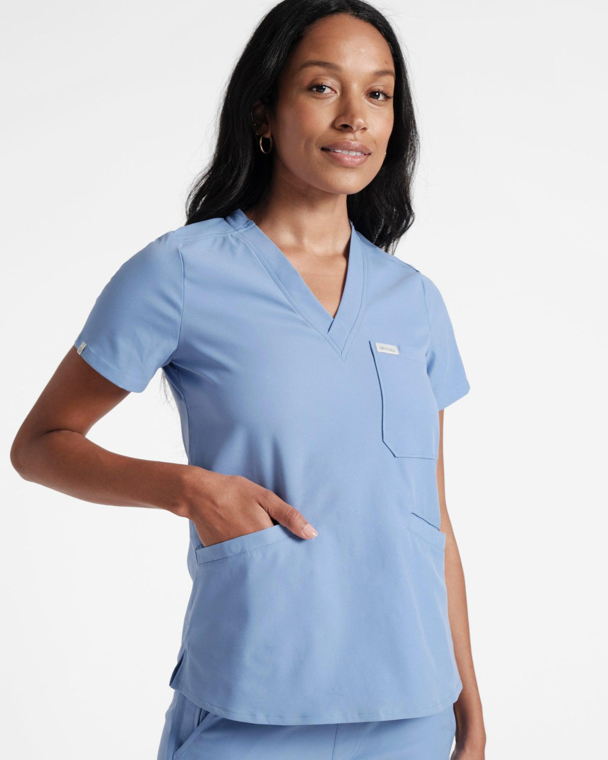 Woman in a v-neck light blue scrub shirt with a chest pocket and two front pockets.