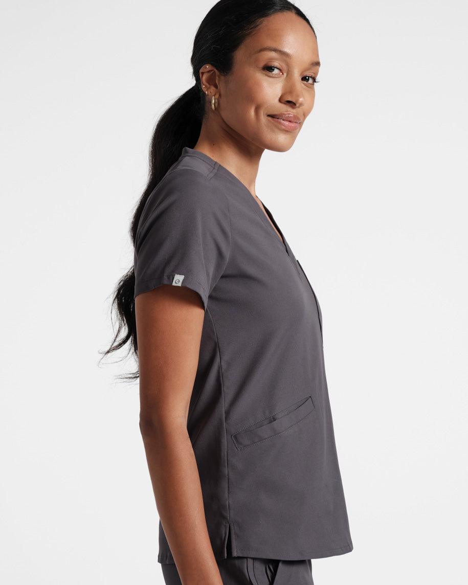 Side view of woman in dark grey scrub top with front pockets and a small fabric logo label wrapped around the sleeve hem. 