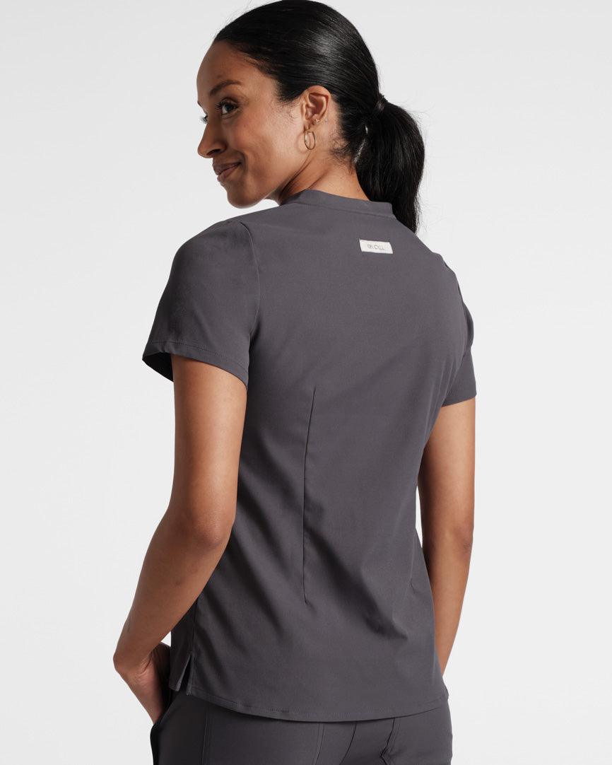 Back side view of woman in dark grey scrub shirt with dart detailing and On Call fabric label under the neck seam. 