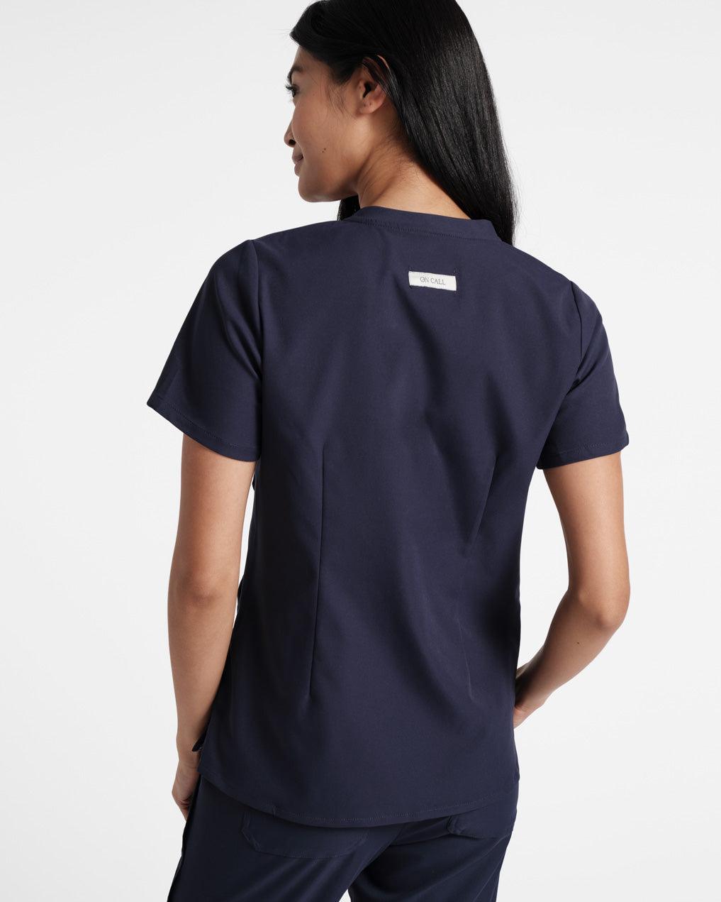 Back of woman wearing a navy scrub shirt with dart detailing and an On Call fabric label sewn under the neck seam. 
