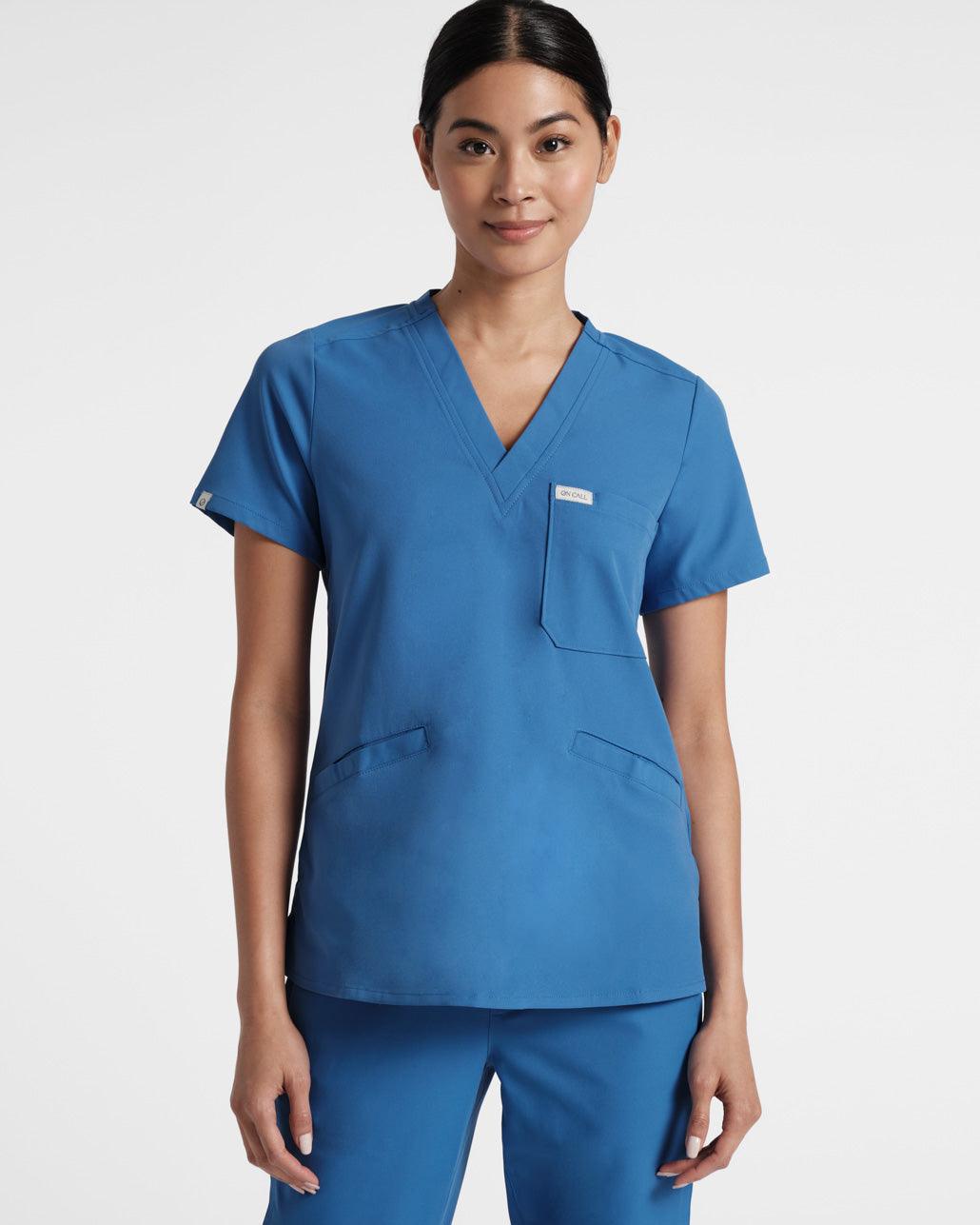 Woman wearing a bright blue v-neck scrub shirt with a chest pocket and two front pockets.