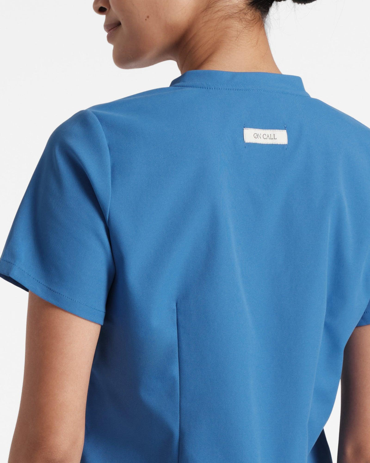Close up view of woman in a bright blue scrub shirt with dart detailing and On Call fabric label sewn on the back under the neck seam.